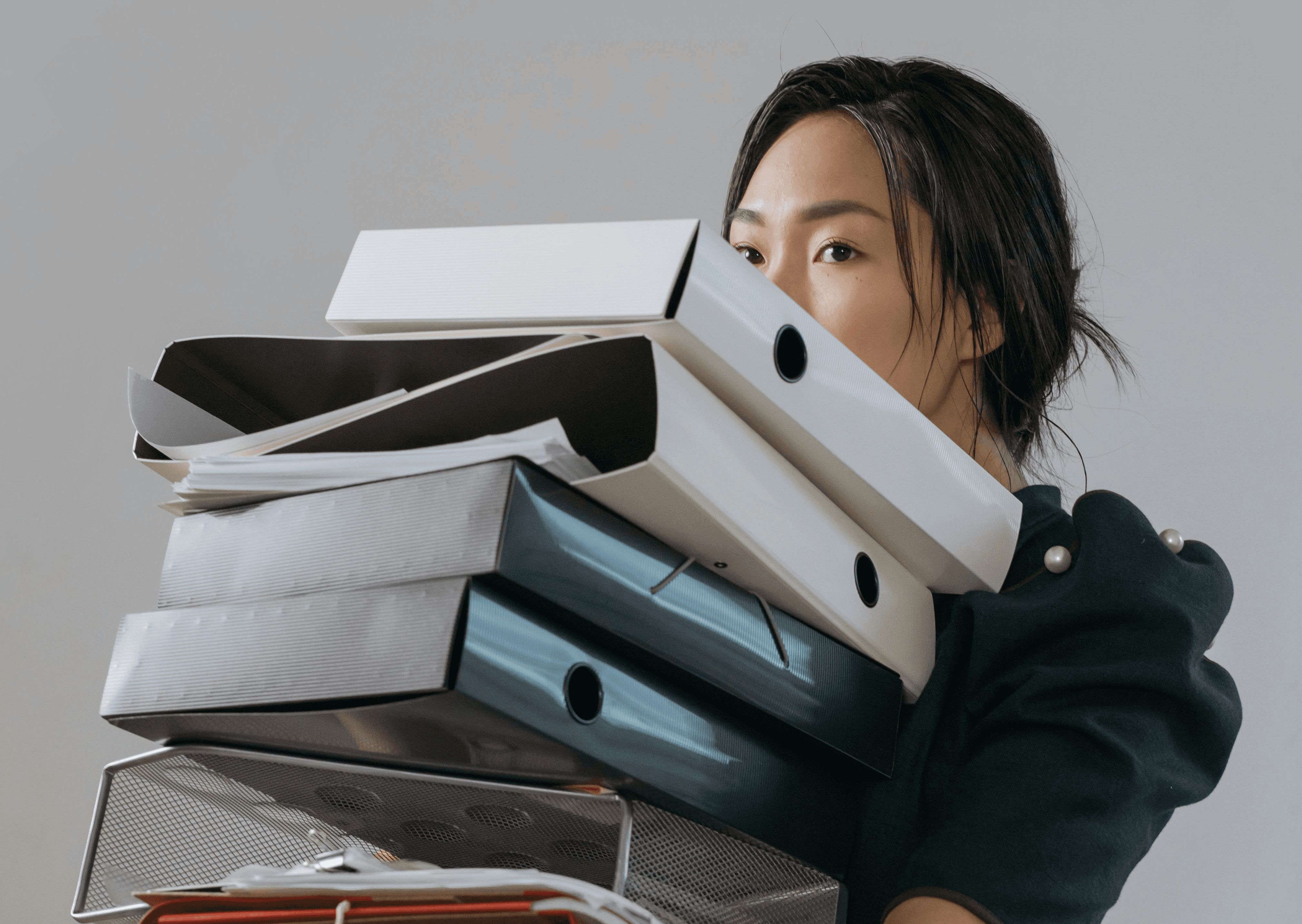 Donna che trasporta una pila di raccoglitori e documenti in ufficio, simbolo di sovraccarico di lavoro e gestione caotica delle attività.