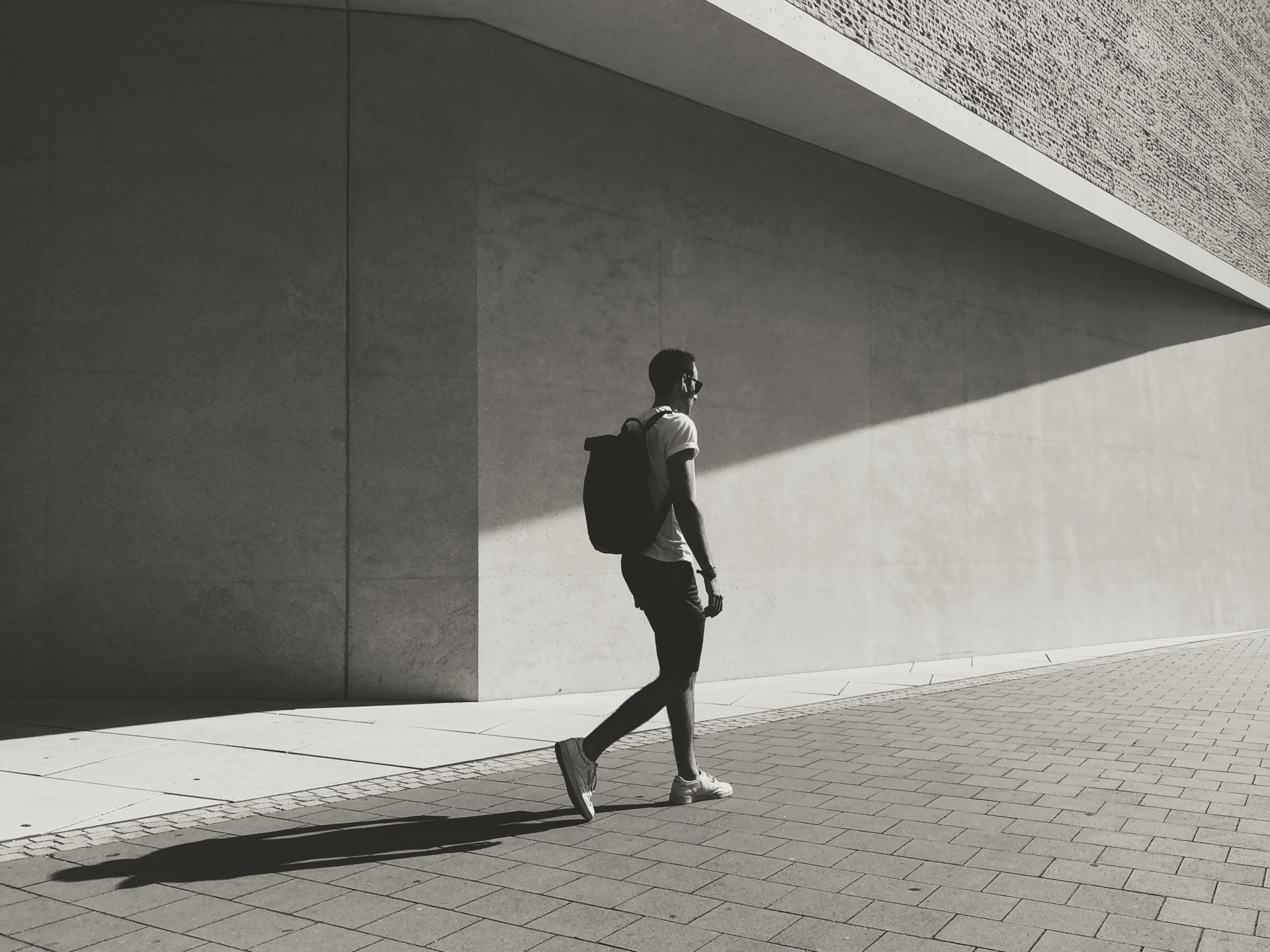 Man walking on brick sidewalk with backpack, passing by large concrete building with sunlight casting shadow.