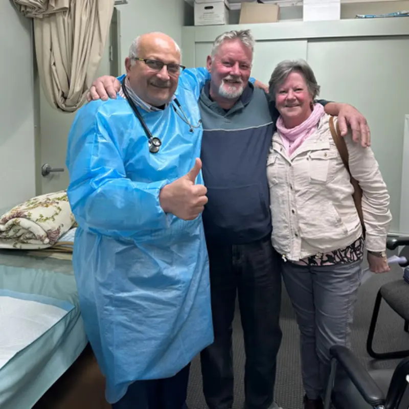 Dr. Ashraf Saddik giving a thumbs-up standing beside an adult couple inside a Coburg clinic examination room.
