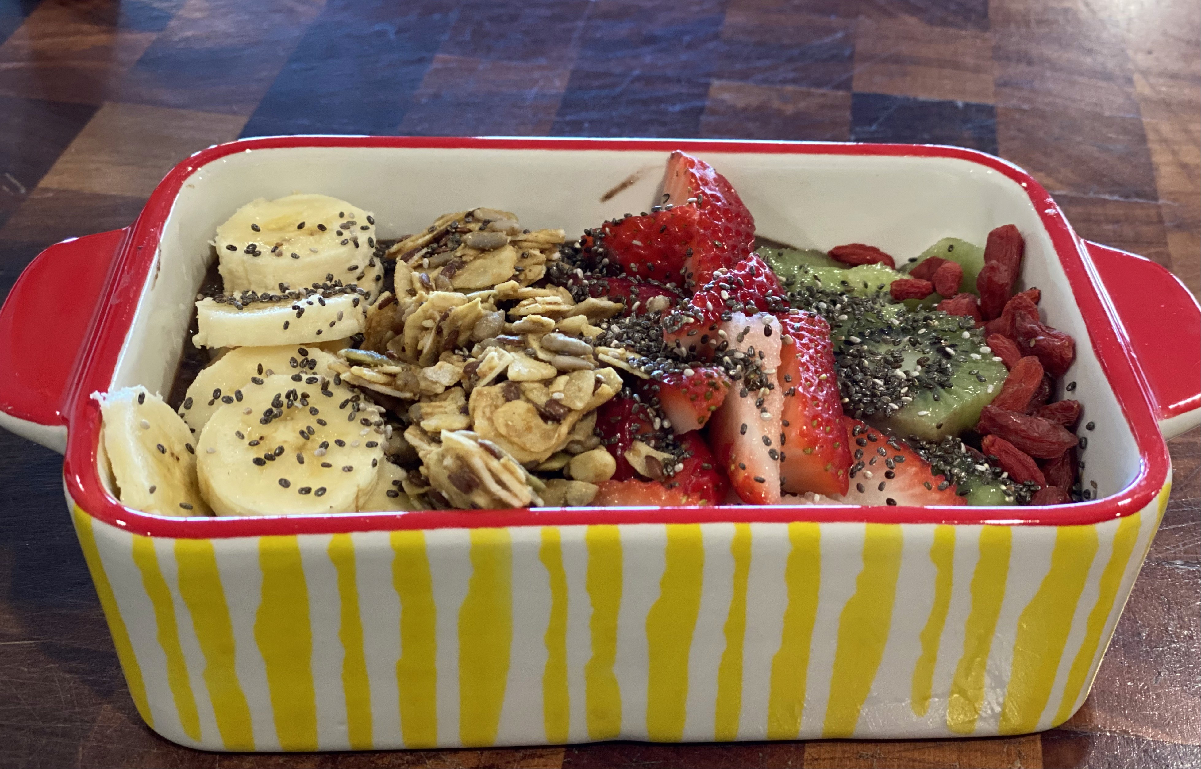 A vibrant breakfast bowl on a wooden table is filled with sliced bananas, strawberries, kiwi, granola, chia seeds, and goji berries, inside a striped yellow and white ceramic dish with red handles.
