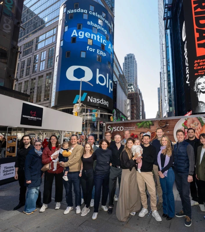 A group of people smiling and posing in Times Square, New York City. Behind them, a large digital billboard displays the Nasdaq logo and a company announcement about AI. The scene conveys excitement and celebration.