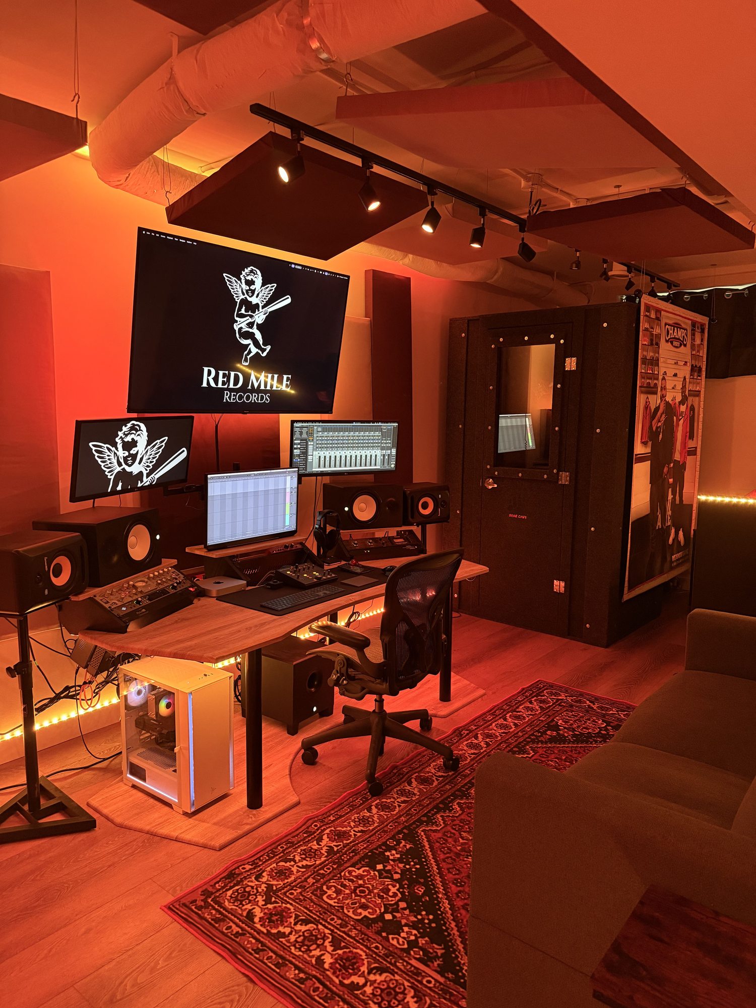 Recording studio control room at Red Mile Records in Calgary, featuring a professional music production setup with studio monitors, mixing desk, multiple monitors displaying the record label logo, an acoustic vocal booth, and a comfortable couch.