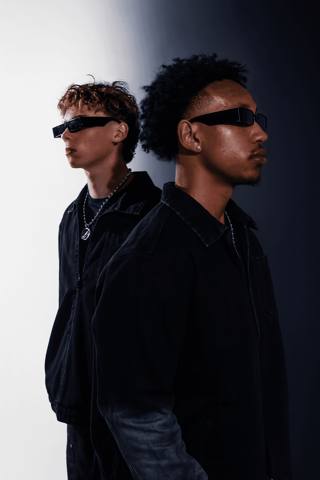 Two men wearing dark outfits and black sunglasses stand against a white-to-navy gradient background