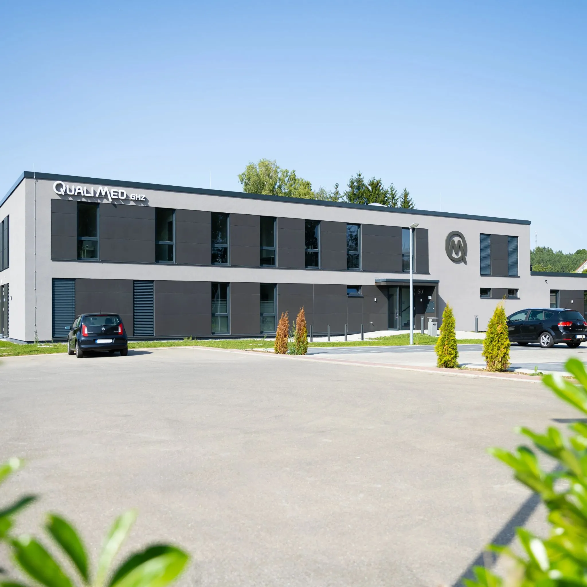 Modernes Grau-schwarzes Bürogebäude mit der Aufschrift QUALIMED GHZ und einem geschwungenen Q-Logo, zwei dunkle Autos und Grünpflanzen vor hellem Himmel.