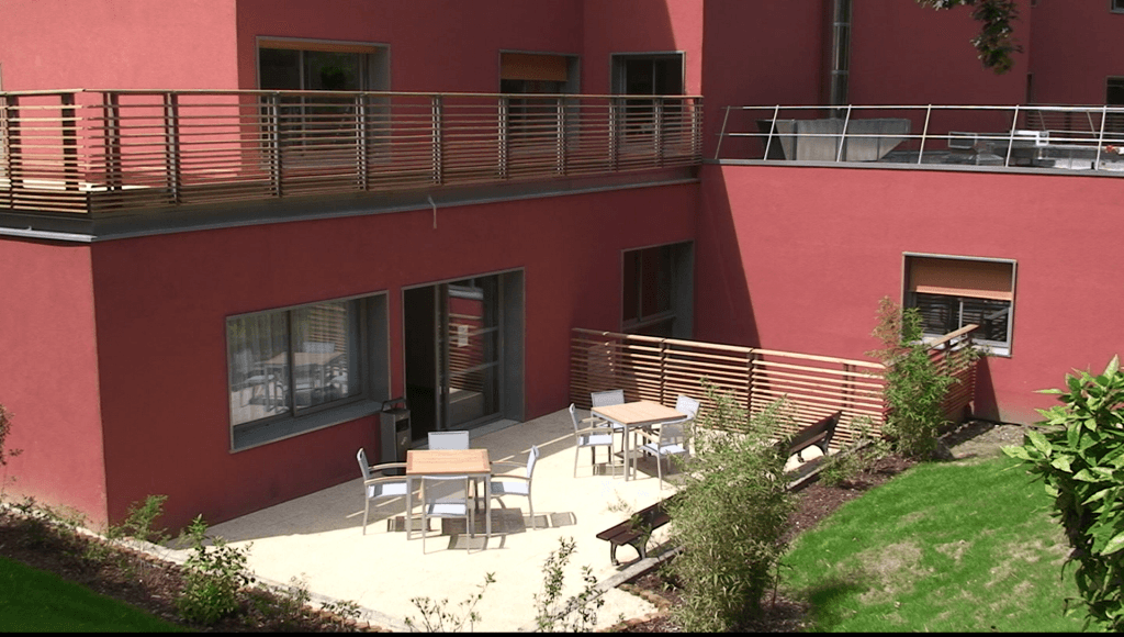 Burgundy-colored residential building with outdoor terrace, wooden decking, dining furniture, green lawn, and bright natural lighting