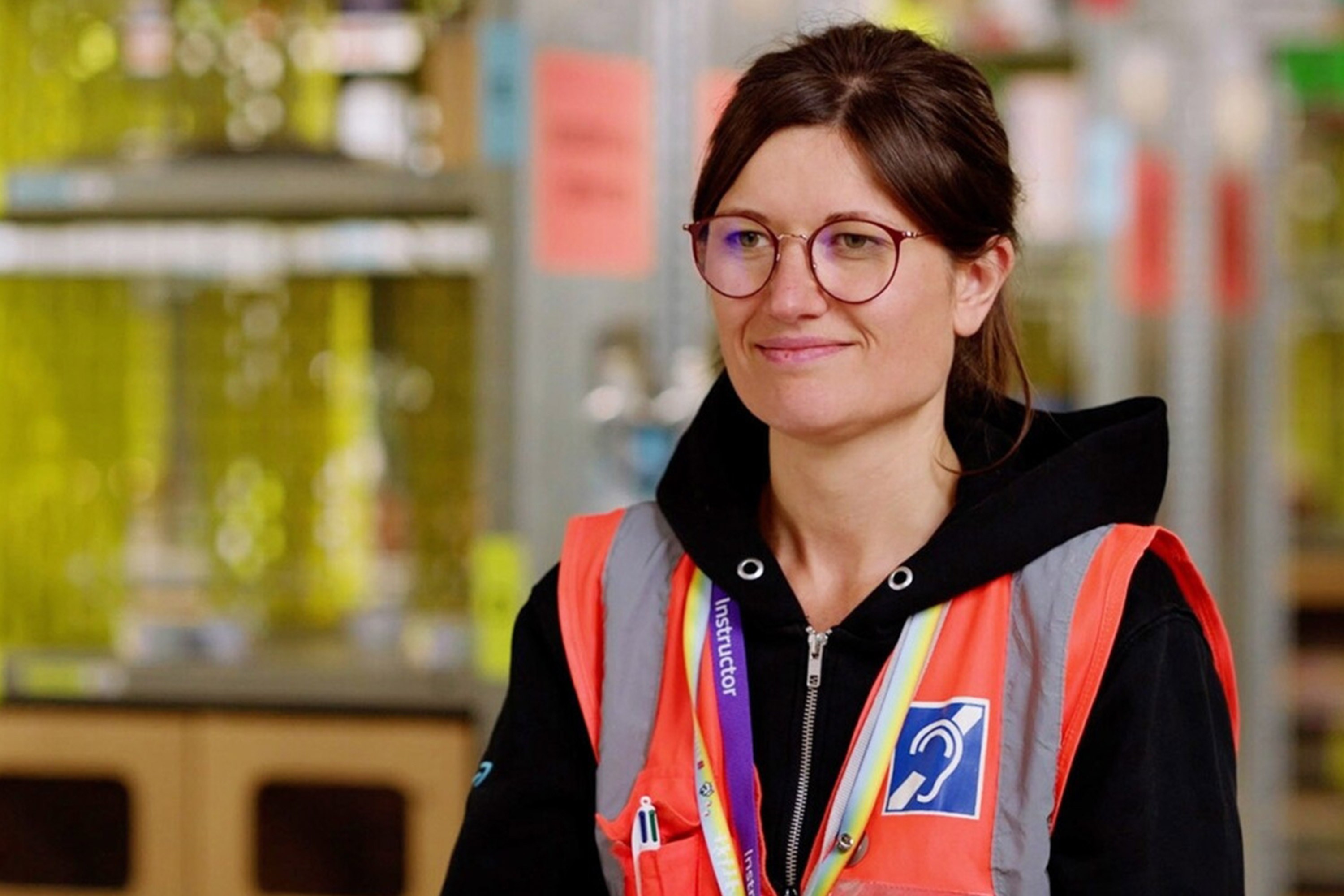 A woman wearing orange safety vest with a symbol representing "deaf" patched on. She is smiling and in the background out of focus is a warehouse setting.
