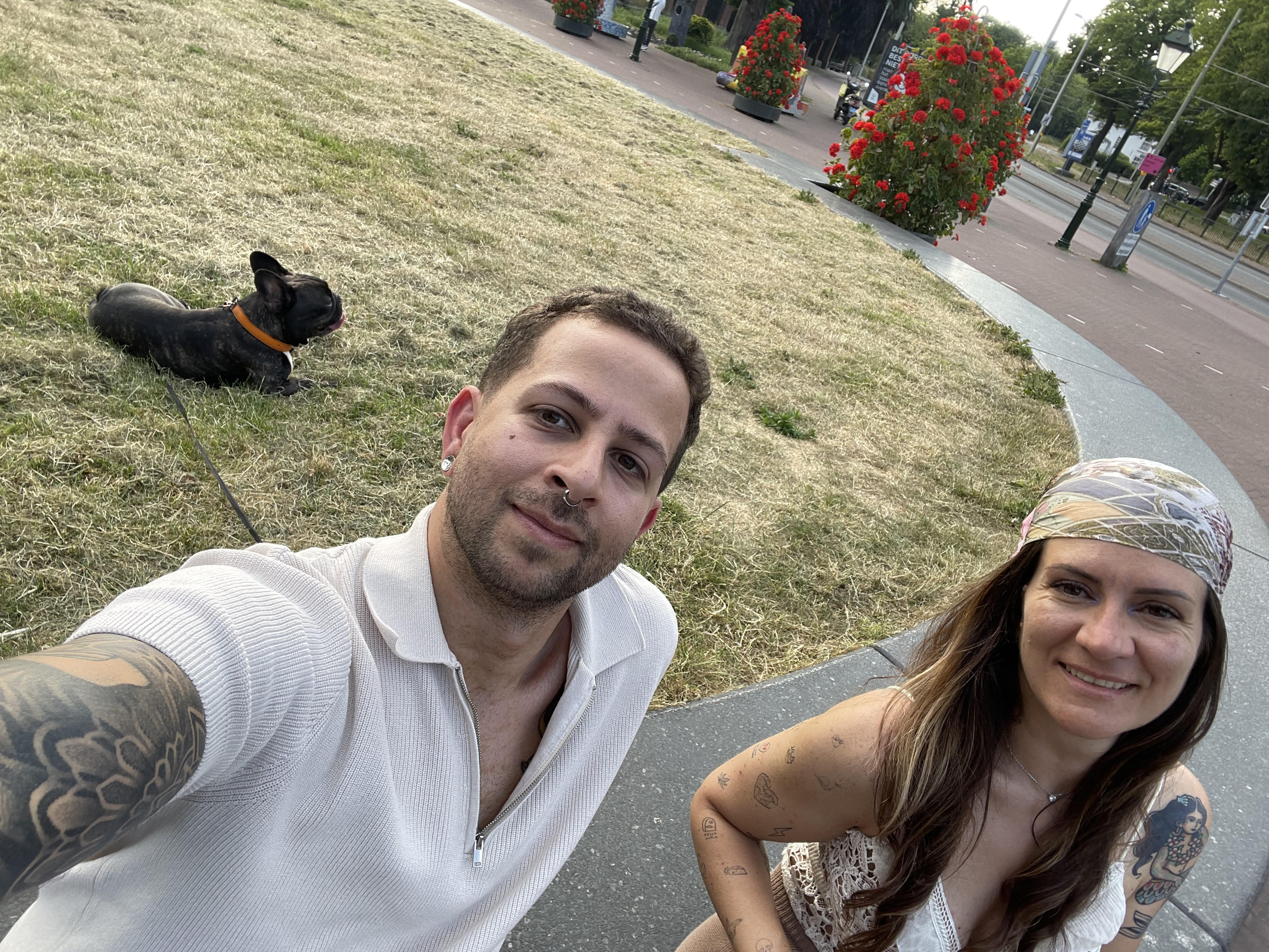 A couple taking a selfie in a park. David, with tattoos and a nose ring, smiles next to his girlfriend in a headscarf. A French Bulldog with an orange collar rests on the grass in the background.