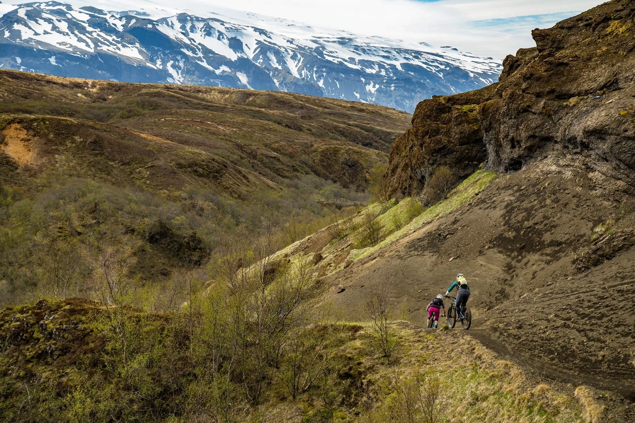 Two cyclists riding a trail below rocky hills and snowy mountains.
