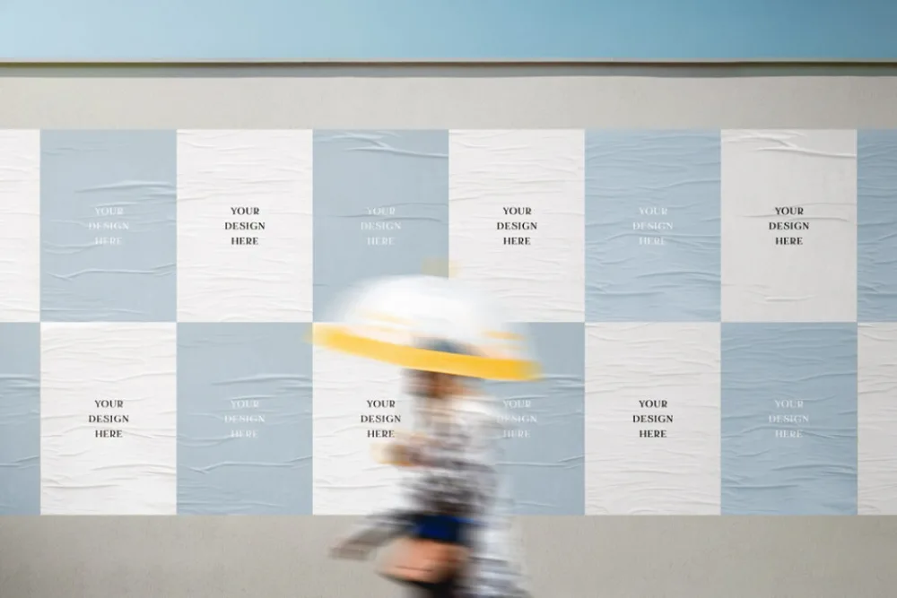 Poster wall mockup featuring alternating blue and white wrinkled paper textures. A blurred figure wearing a yellow hat passes in front, adding motion to the scene.