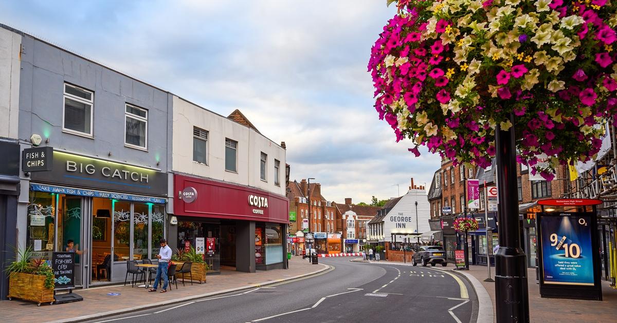 A smiling man with curly hair is sitting at a desk, looking at the camera, with a laptop and a lamp in the background.