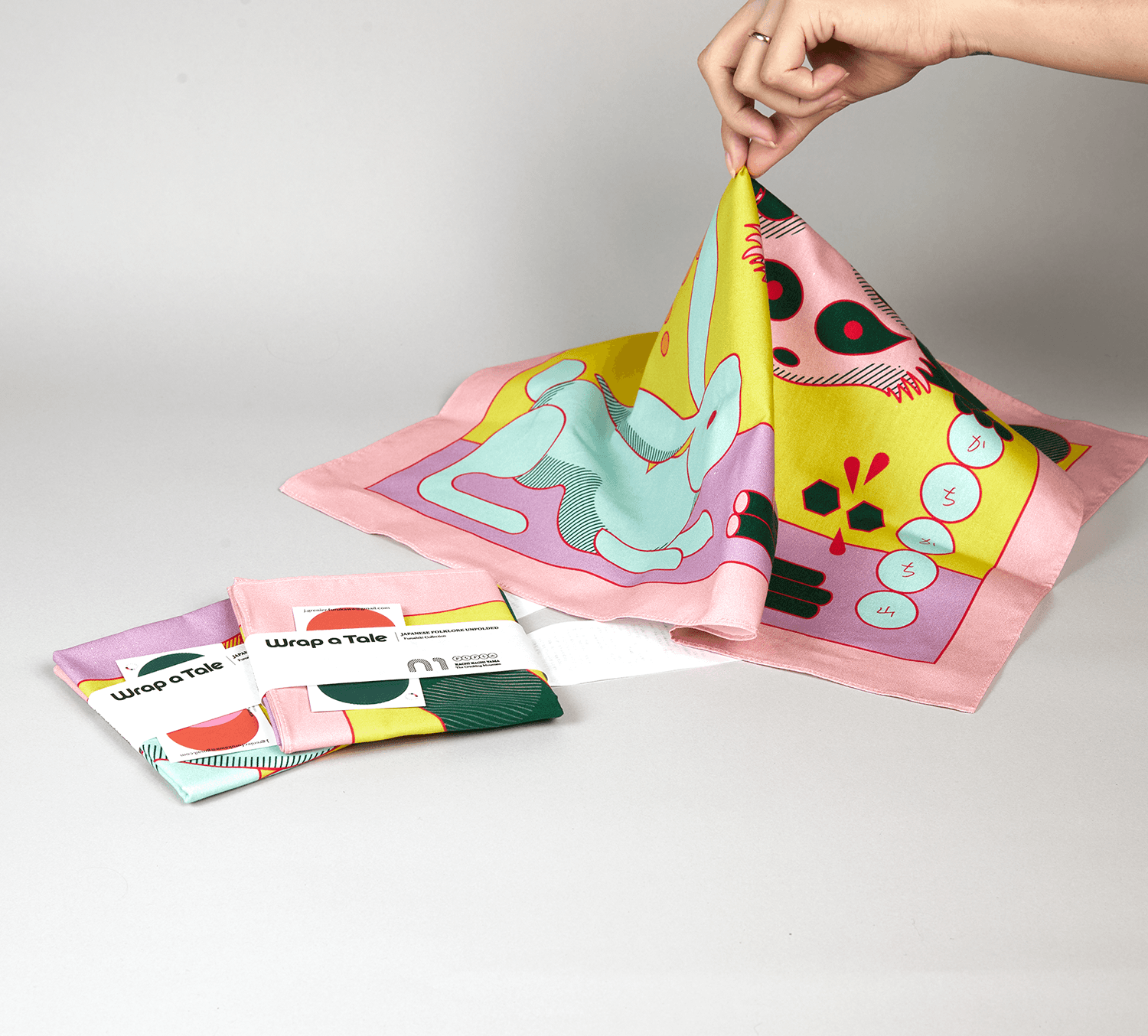 Wrap a Tale furoshiki designed by Josephine Grenier being lifted to reveal its colorful Japanese folklore illustration, with packaged cloths displayed beside it.