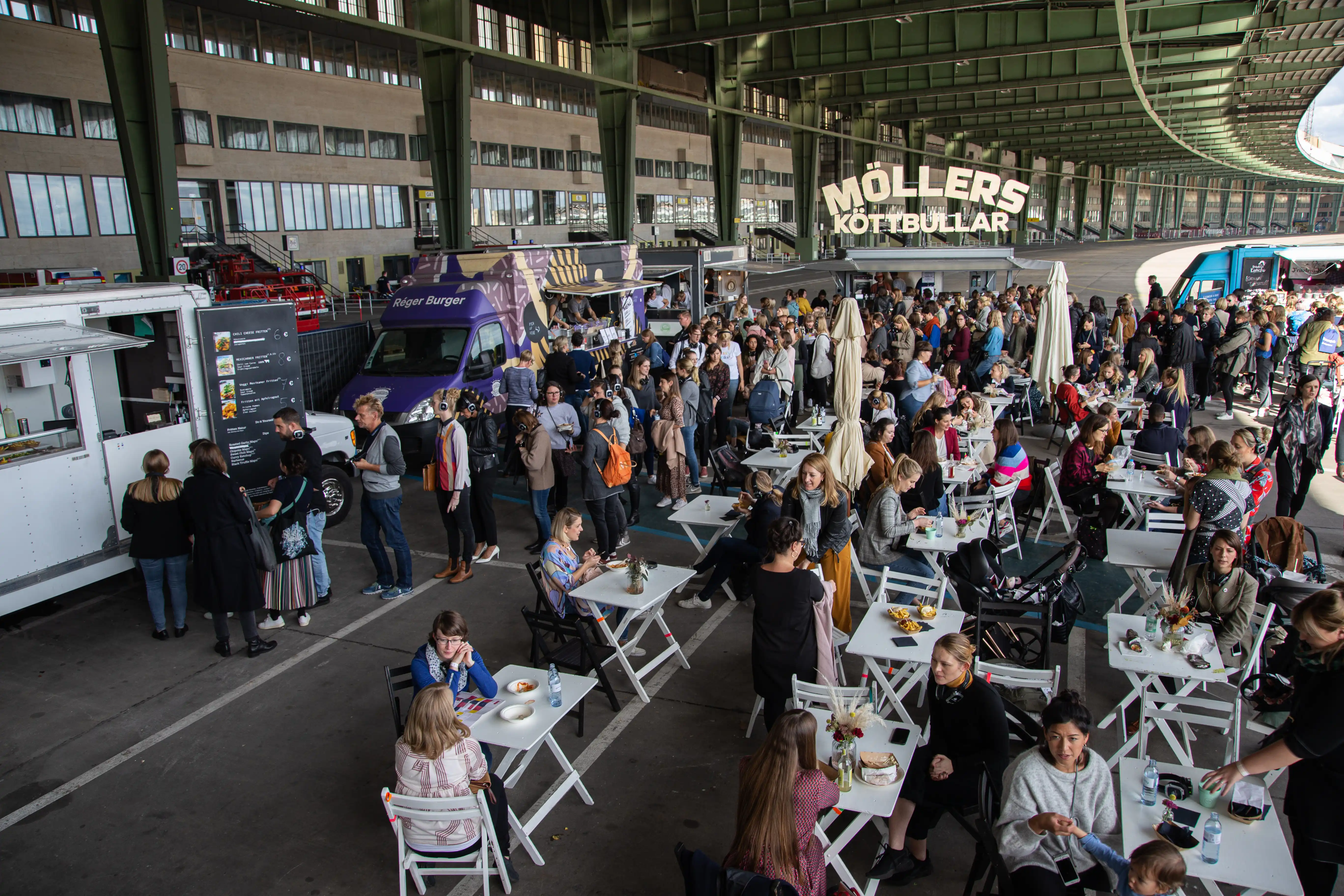 Food truck area with white bistro tables and crowds of visitors under the wide hangar roof at Female Future Force Day.
