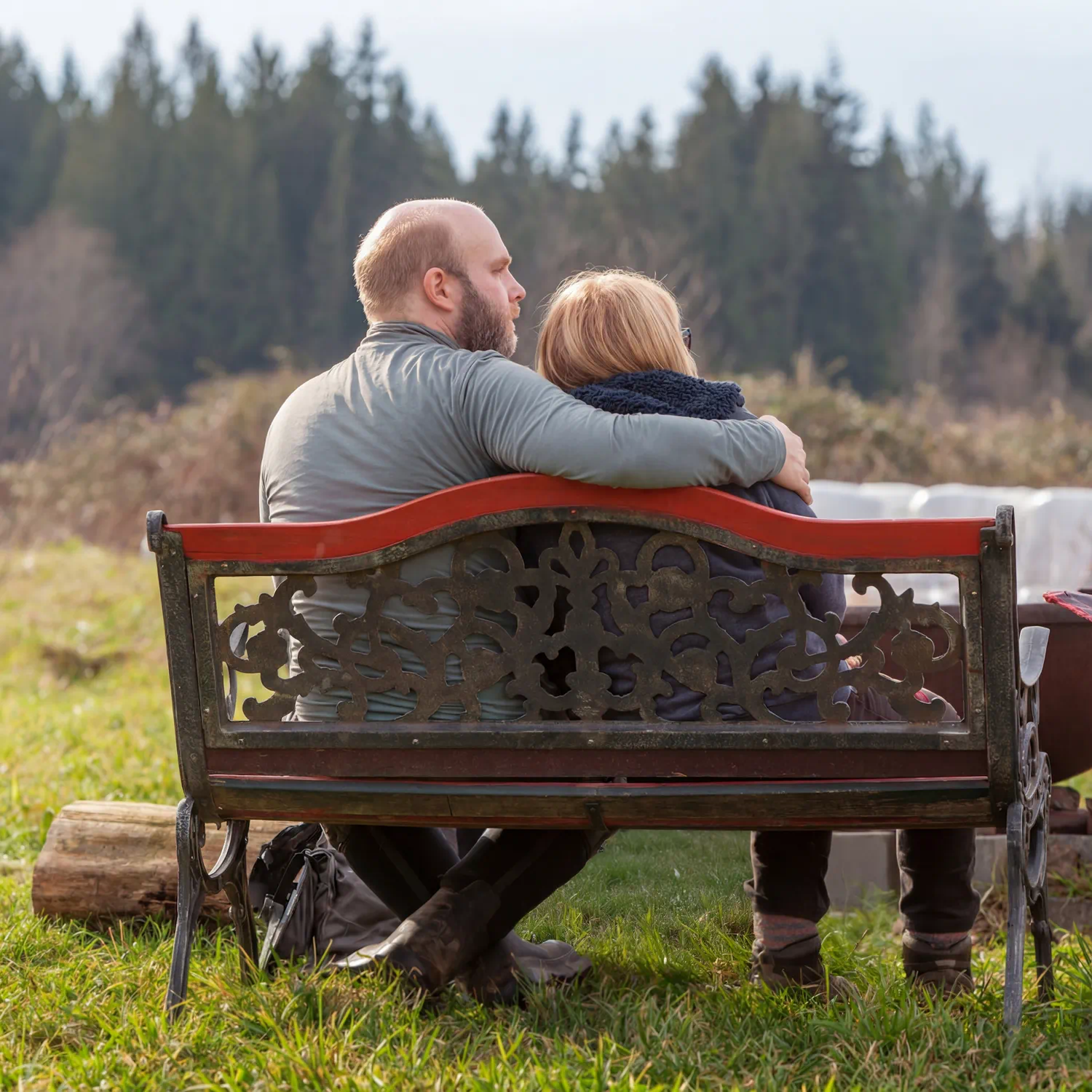 Couple sitting close together on a red metal bench overlooking an open field with forested hills in the distance.