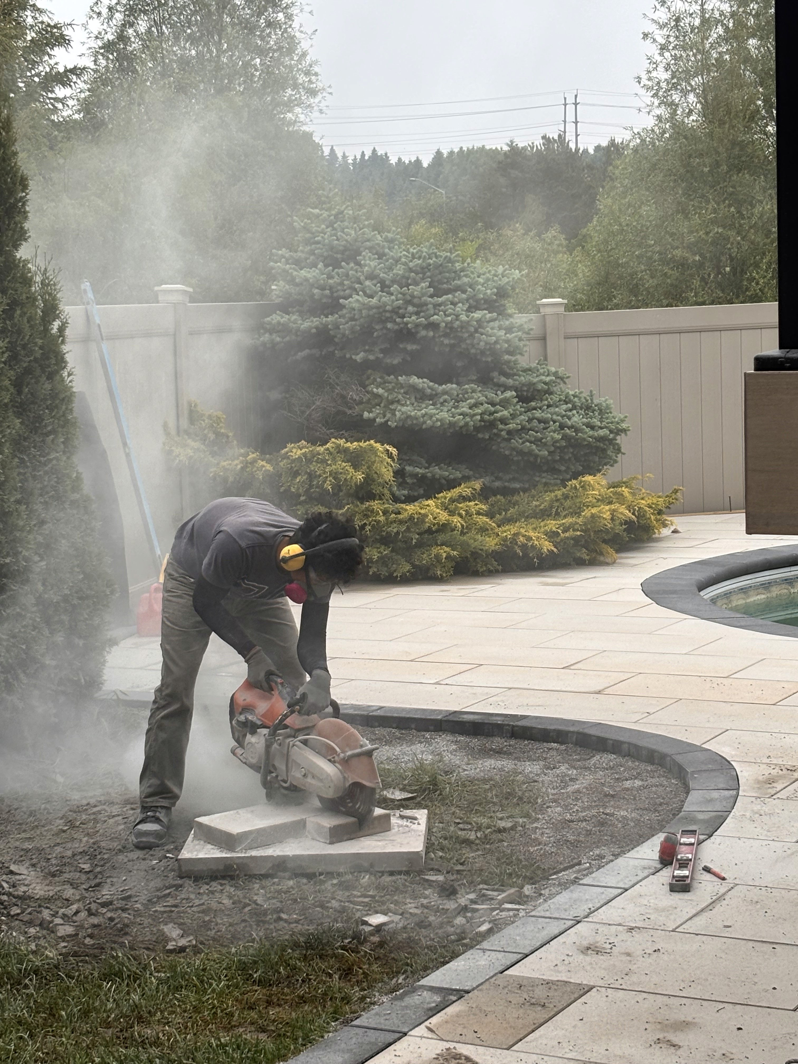 A person cutting pavers near a pool, surrounded by greenery.