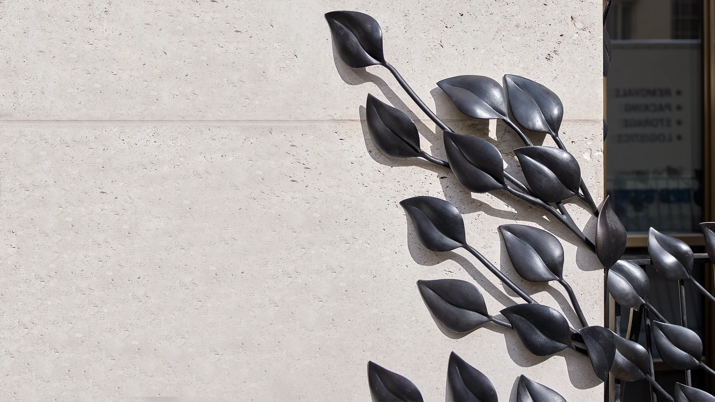 Close-up of hand-forged dark bronze sculptural leaves sprawling across a textured beige limestone wall. This architectural metalwork is an independent objet d’art installation designed by Shaun Leane.