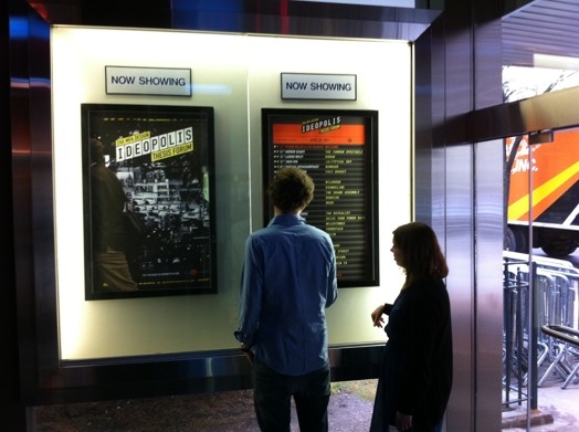 A photo of two people looking at two posters with the title IDEOPOLIS.