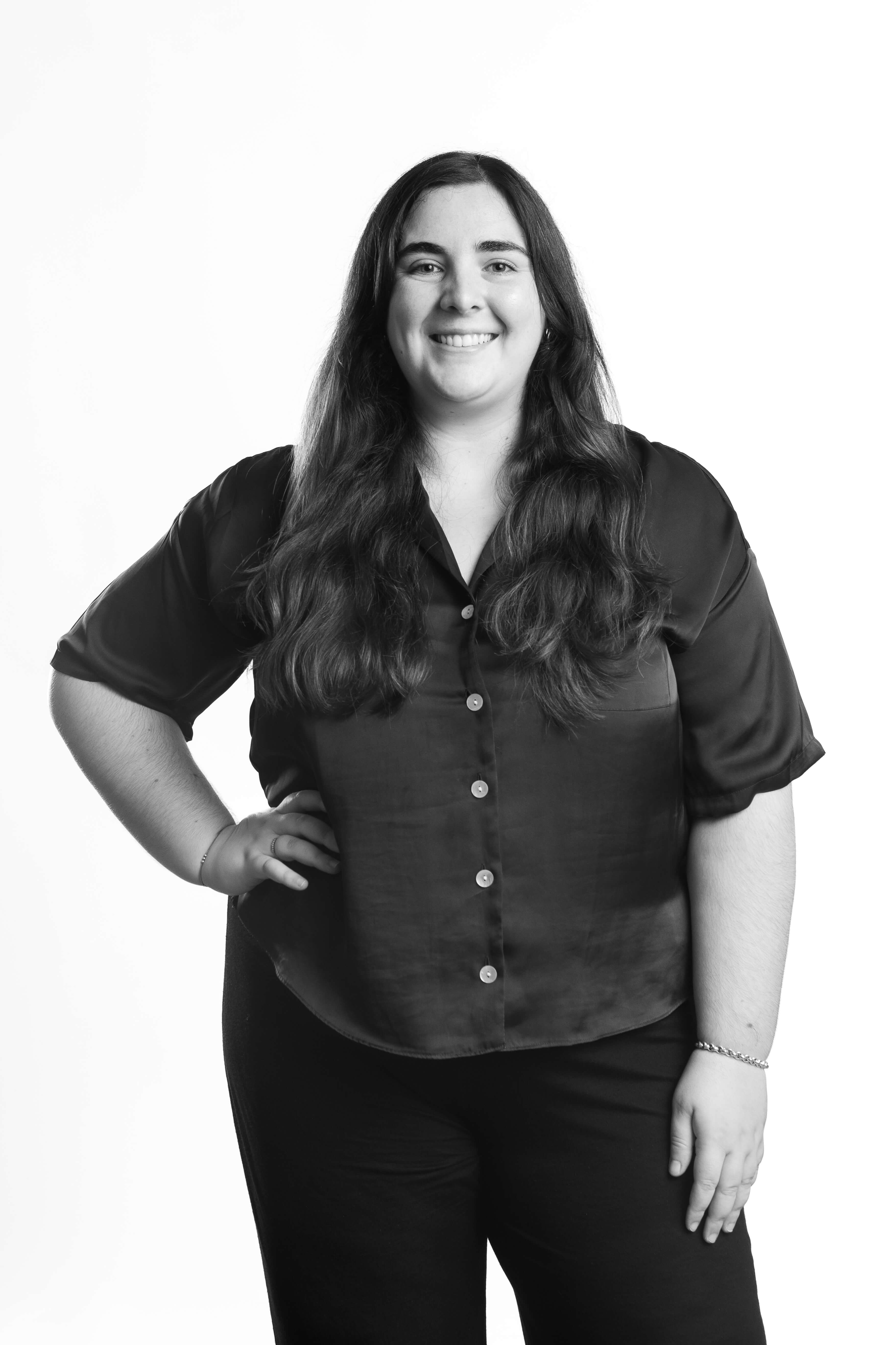 Black and white photo of a smiling woman standing confidently with long hair, wearing a button-up shirt and pants. The background is plain white.