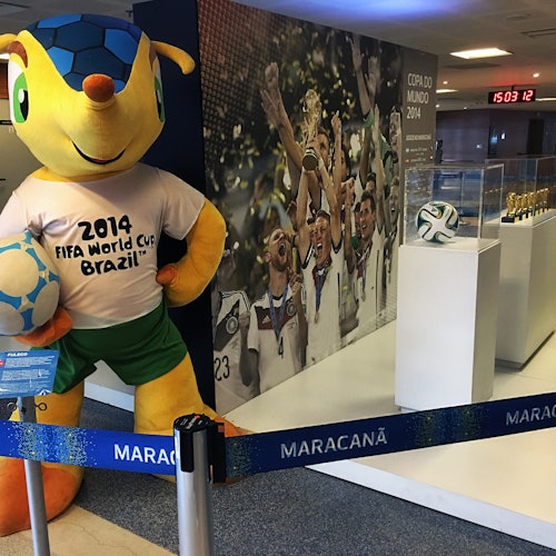Mascota Cupei Mondiale FIFA 2014 stă lângă o expunere de trofee și o fotografie a jucătorilor care sărbătoresc o victorie pe stadionul Maracanã.