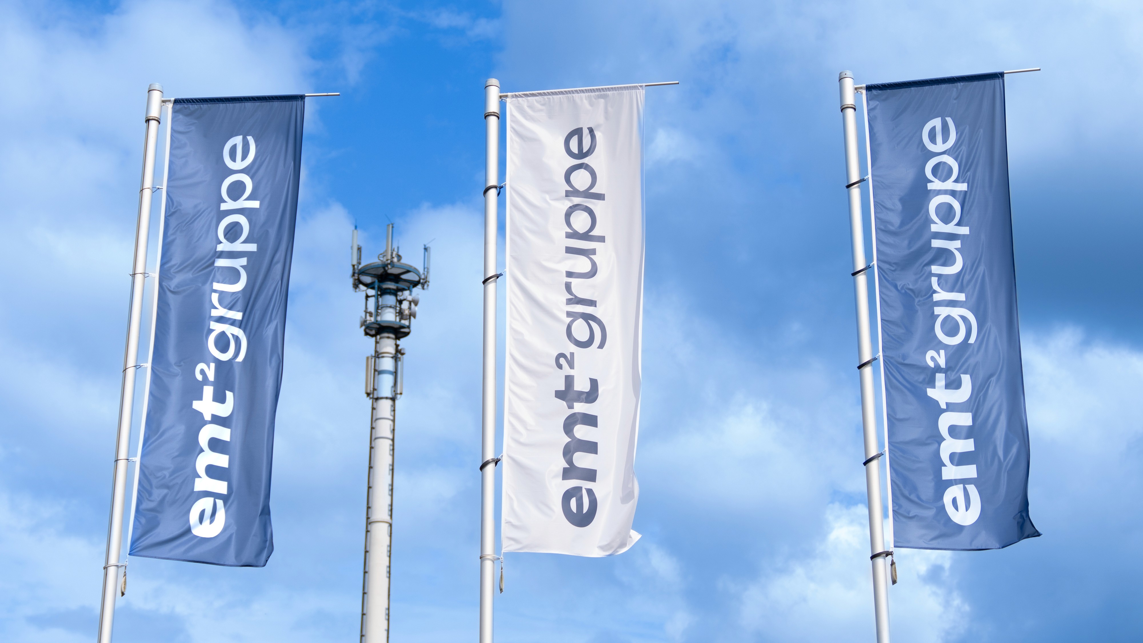 emt²gruppe Fahnen im Freien. Firmenlogo auf blau und weiß vor blauem Himmel.