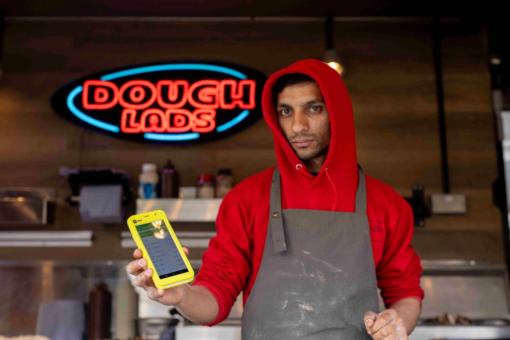 a business owner holds a Teya card machine, he's wearing a grey apron and a red hoodie
