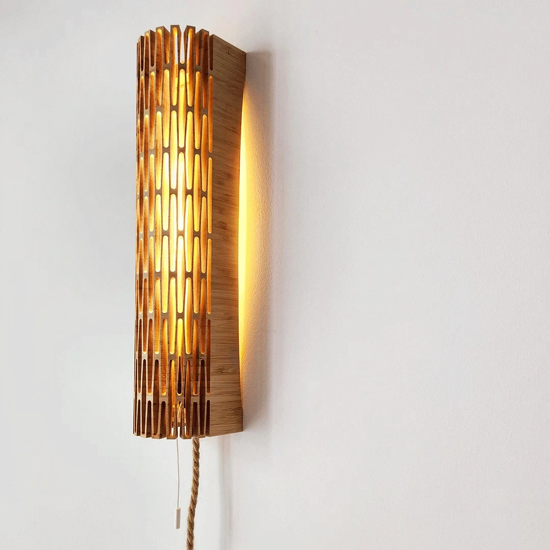 A vertical wall-mounted sconce made of slatted light wood with warm light glowing through the cutouts, featuring a pull string and rope cord on a white wall.