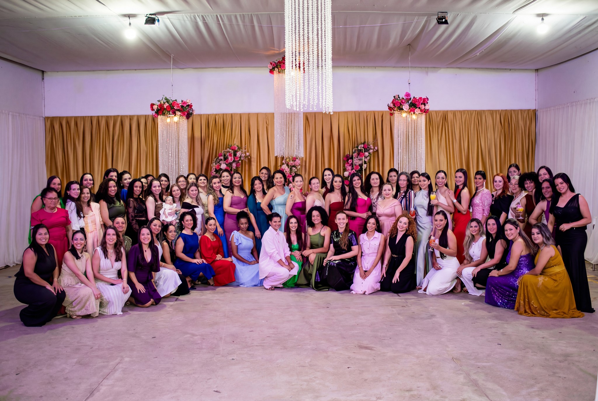 Um grande grupo de mulheres participantes do Evento Conexão Delas, vestidas com trajes de festa variados, posa para uma foto em grupo no salão de eventos. O espaço está decorado com cortinas douradas, arranjos florais e lustres pendentes.