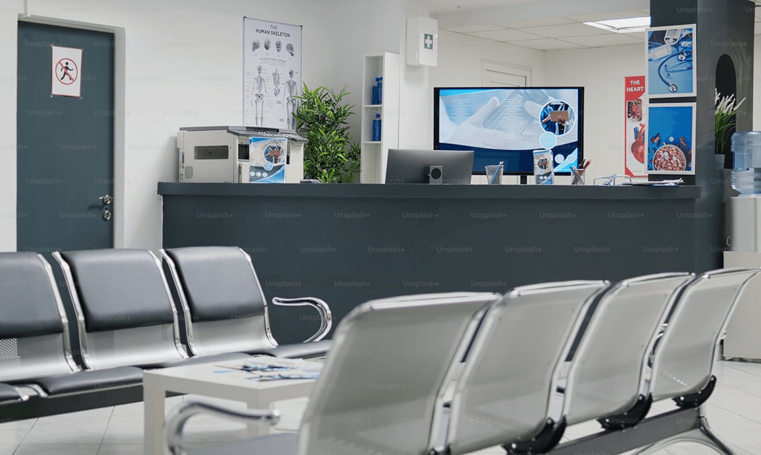 The image depicts a modern medical clinic waiting area with sleek metal chairs, a reception desk featuring a computer monitor displaying medical graphics, and informational posters about anatomy and health on the walls, creating a professional and welcoming atmosphere.