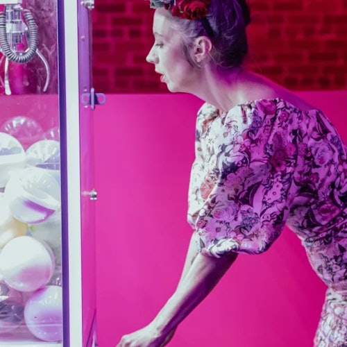 A woman with a floral dress and flower crown operates a claw machine filled with white balls, set against a pink background.