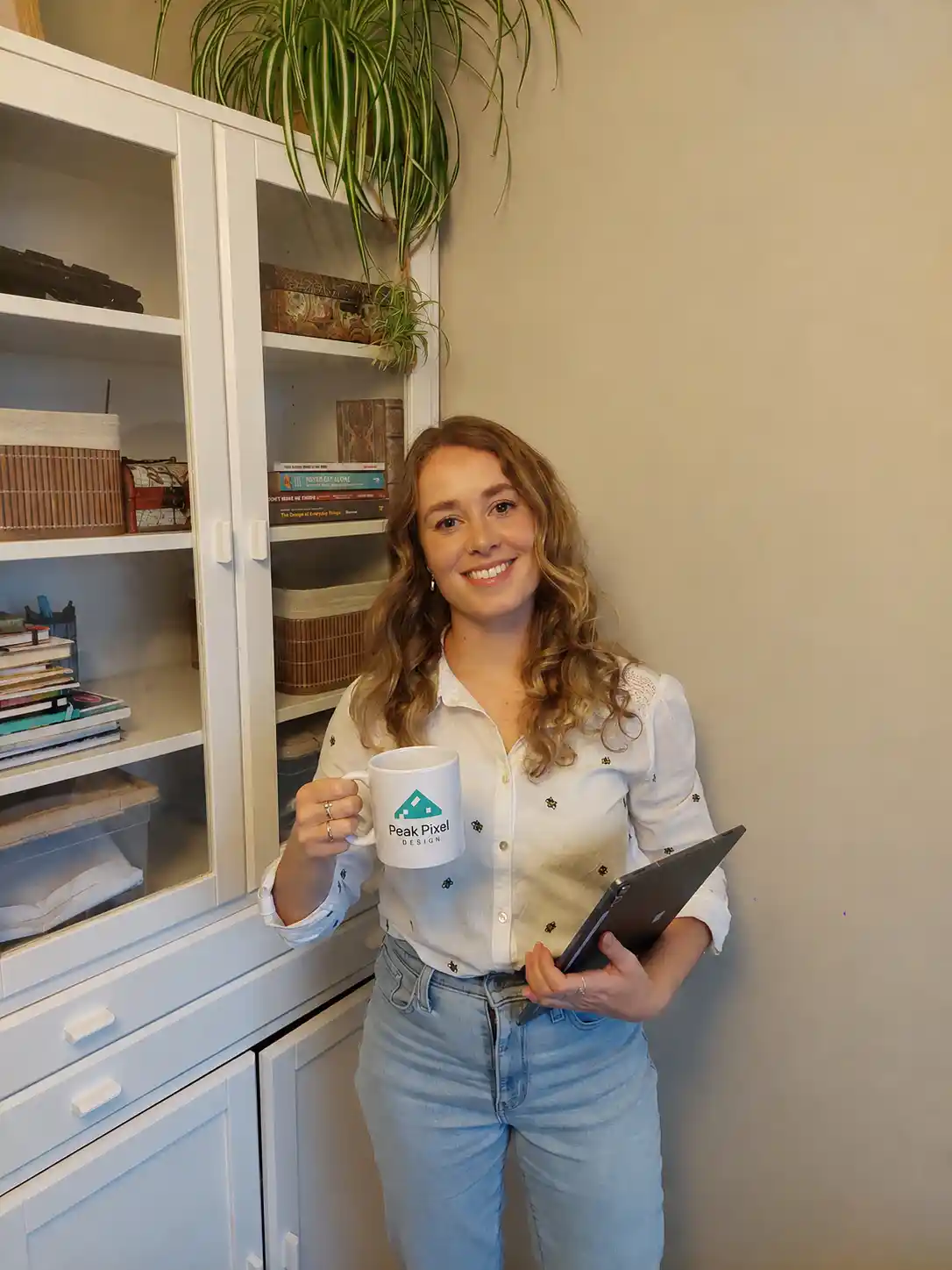 A portrait of Peak Pixel Design founder Yosta in her home office, holding a branded coffee mug and a tablet, smiling and inviting potential clients to work with her.