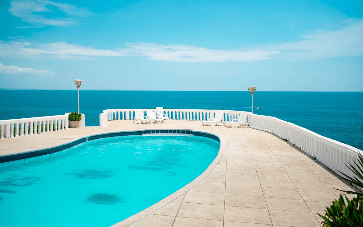 Aerial view of a luxury infinity pool on a tiled deck with white railing, overlooking a clear blue ocean under a sunny sky.