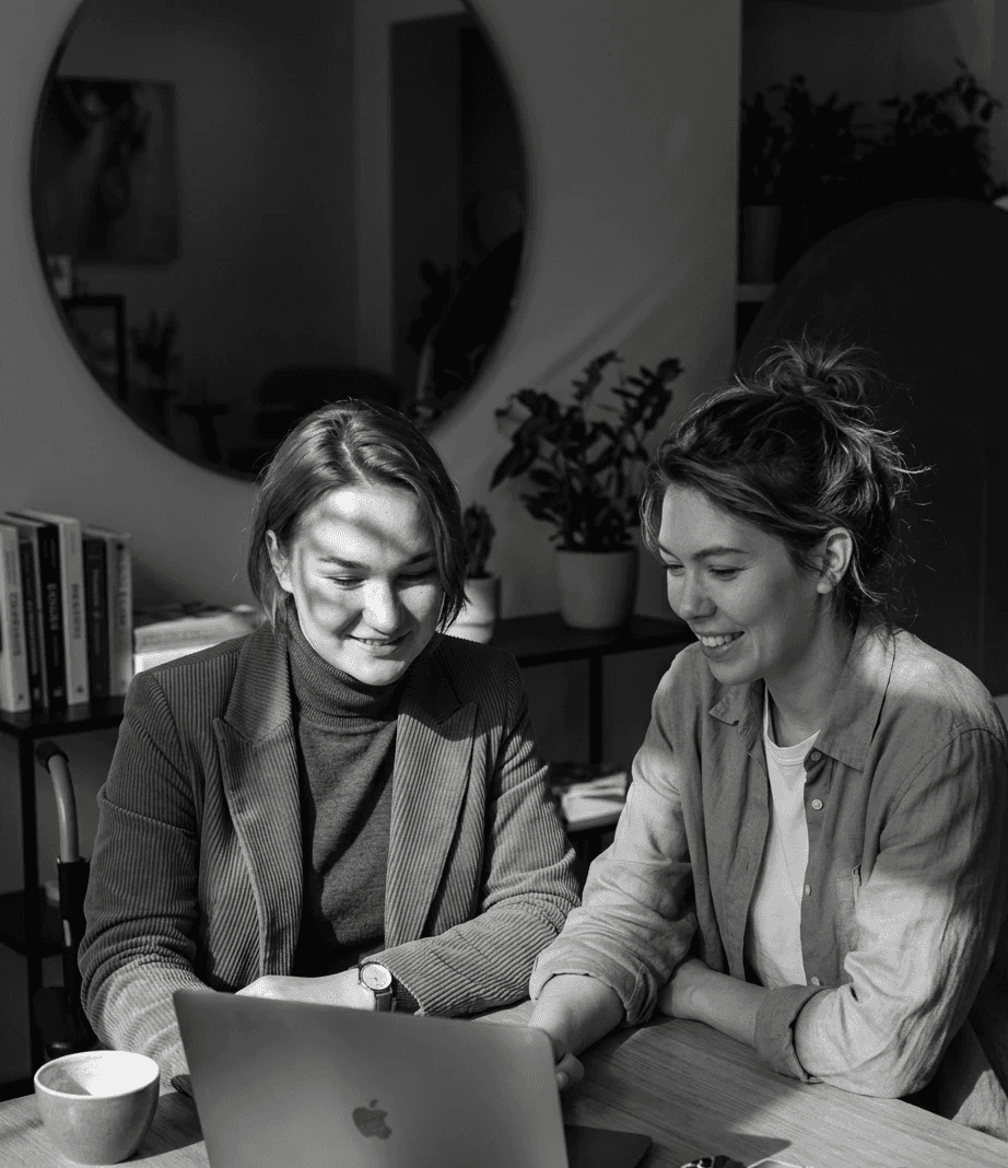 Two women sitting together in a cozy, well-lit workspace, smiling and collaborating over a laptop, with a coffee cup and a bowl on the table.