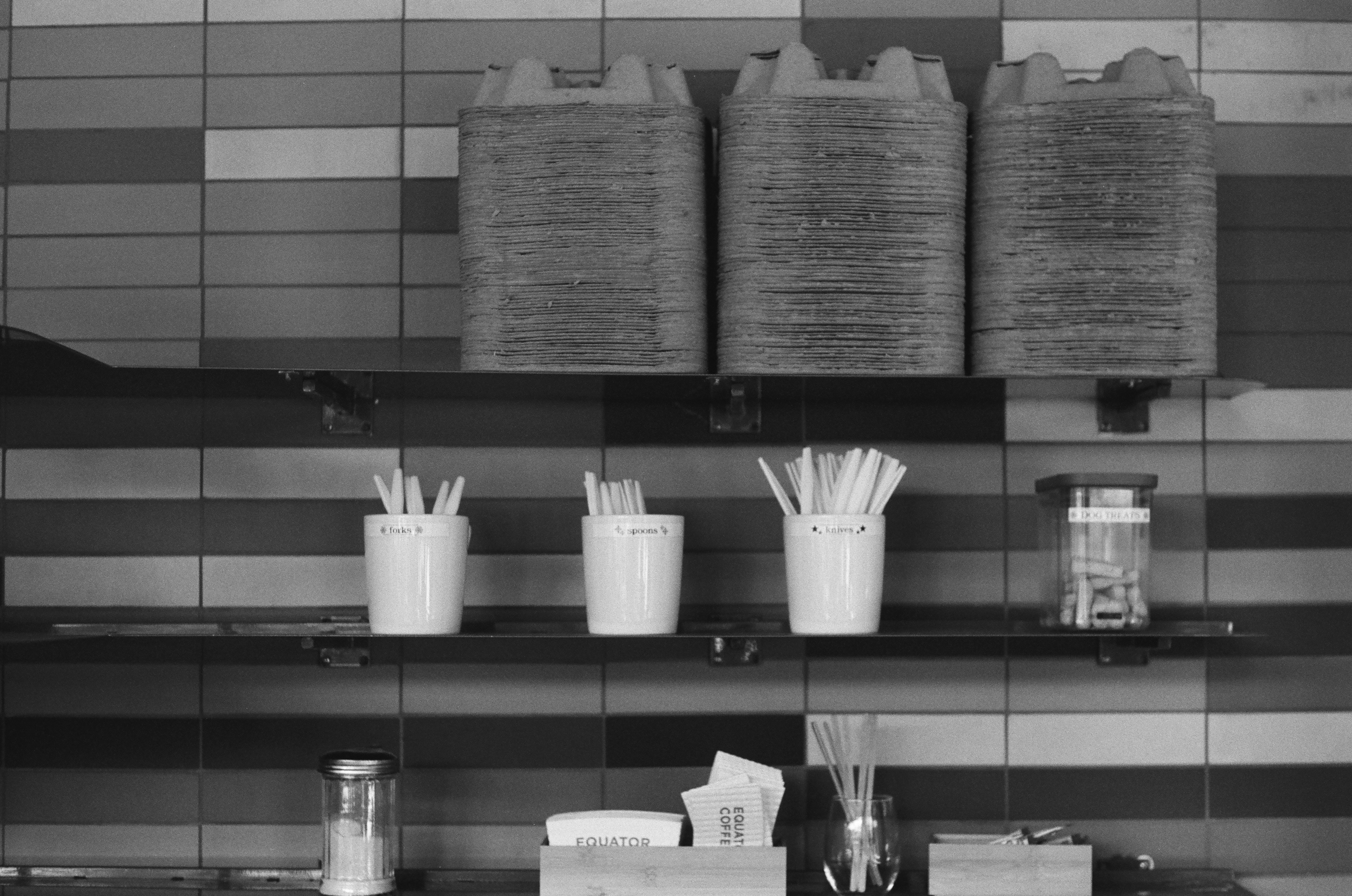 Shelves of takeaway drink carriers and labeled cutlery containers against a tiled wall at Equator Coffees in Culver City.
