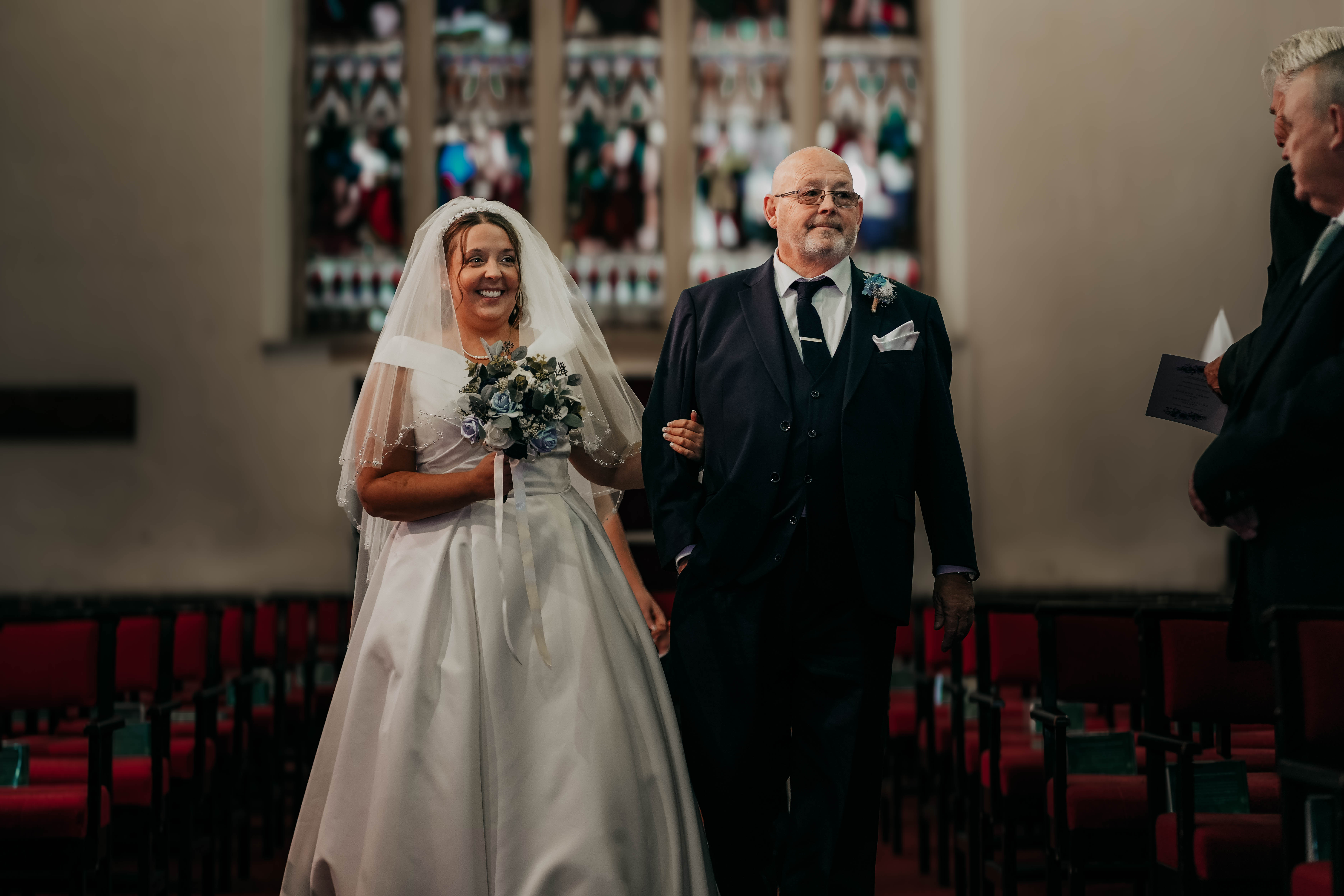 A smiling bride in a white A-line gown and veil holds onto her father's arm as he walks her down the aisle of a church.