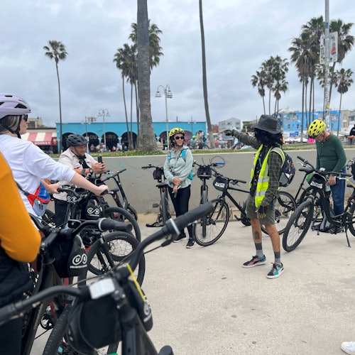 Un grupo de personas con bicicletas se reúne al aire libre, cerca de palmeras y edificios, algunos vistiendo cascos y chalecos reflectantes.