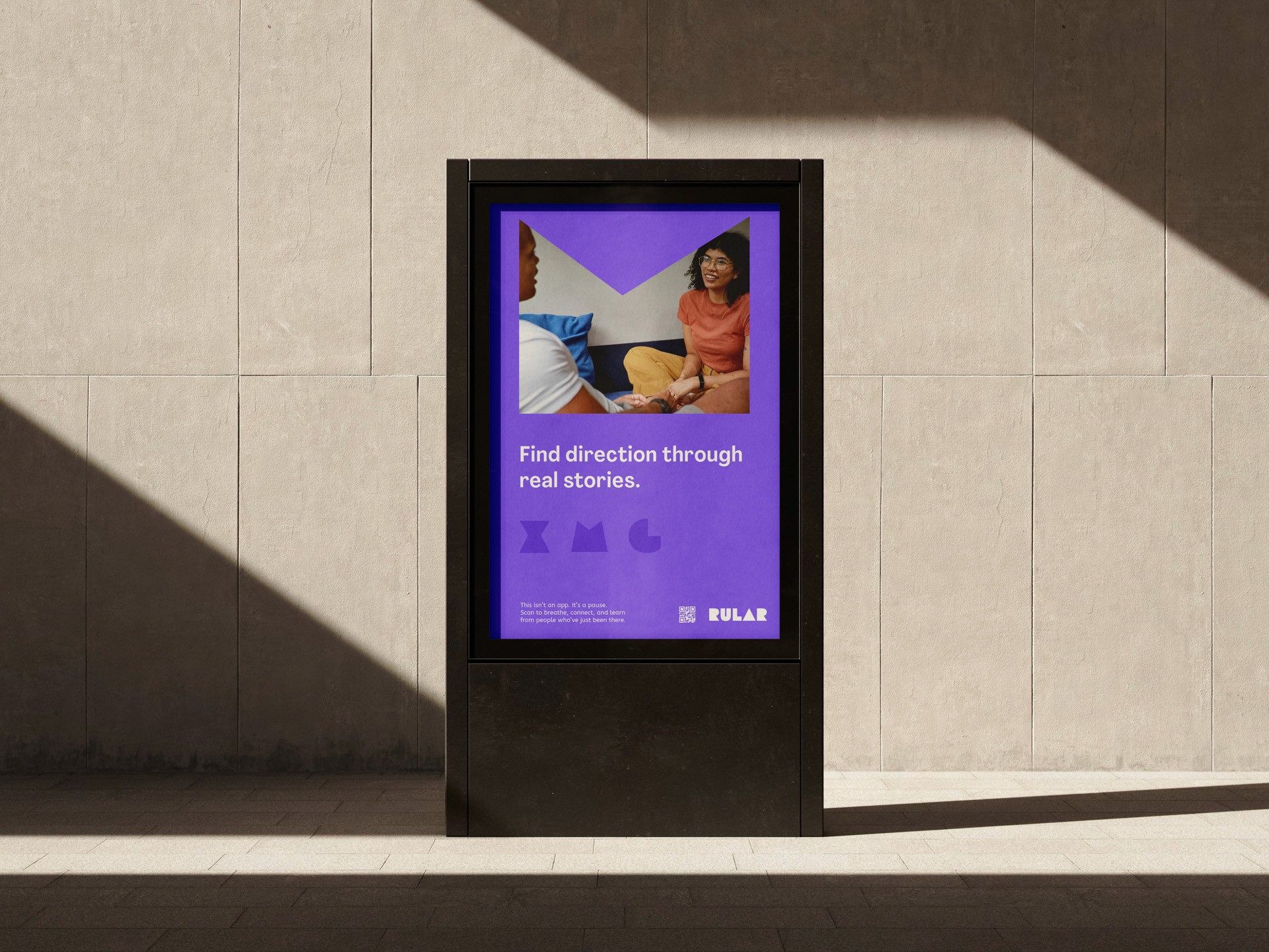 A purple digital display screen mounted on a dark gray concrete pillar shows a person seated on a couch, looking toward the viewer. The screen displays the text "Find direction through real stories." Below the text are three stylized letters: "xMG." To the right of the text is a small logo that says "RULAR." A shadow falls across the wall behind the pillar, partially covering the display.