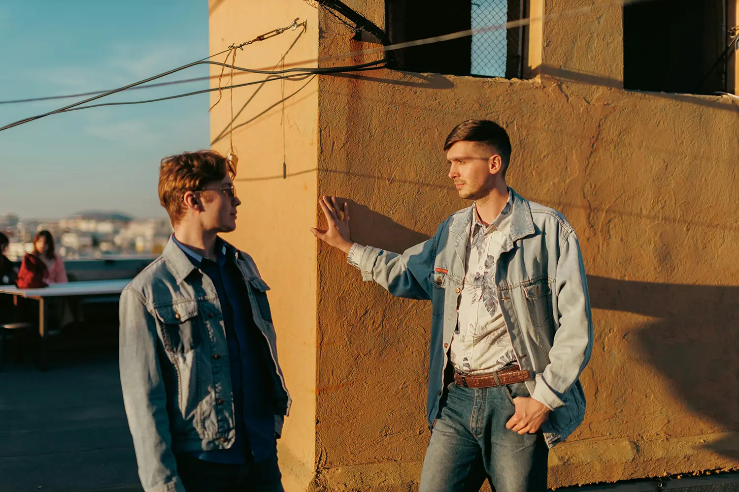 Two young men in denim jackets talk on a rooftop at sunset