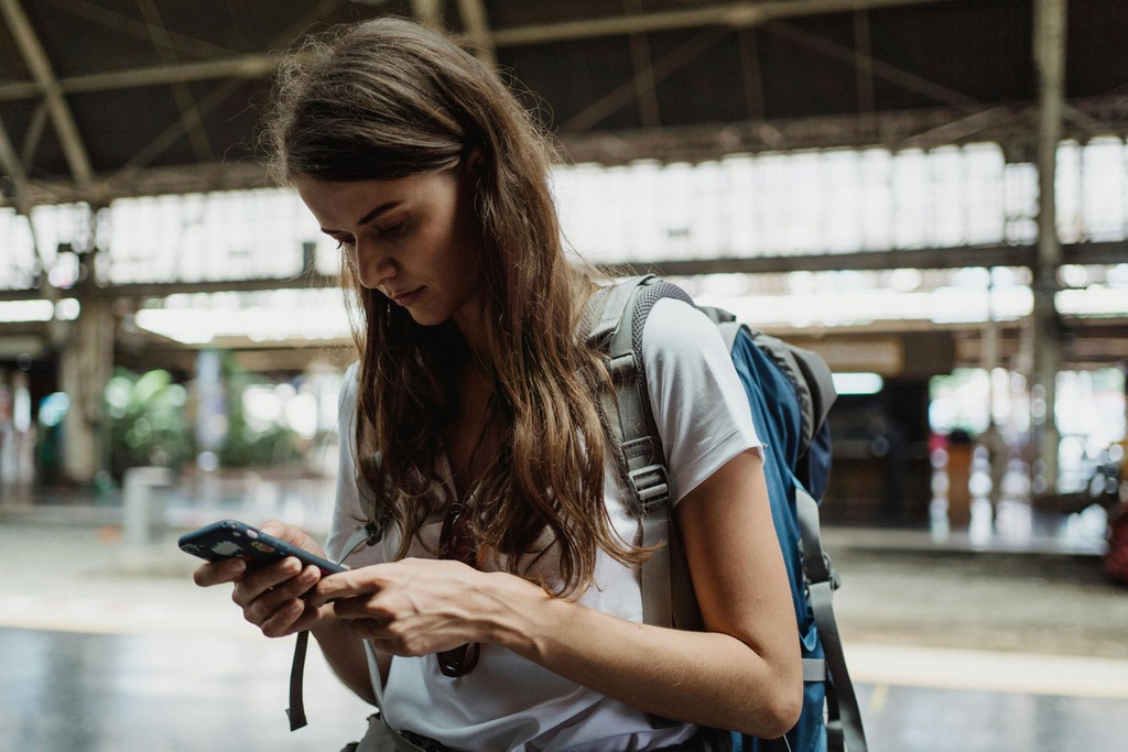 A young woman with a travel backpack looks intently at her smartphone in a busy airport terminal, preparing to activate a digital travel eSIM to avoid high Canadian roaming fees.