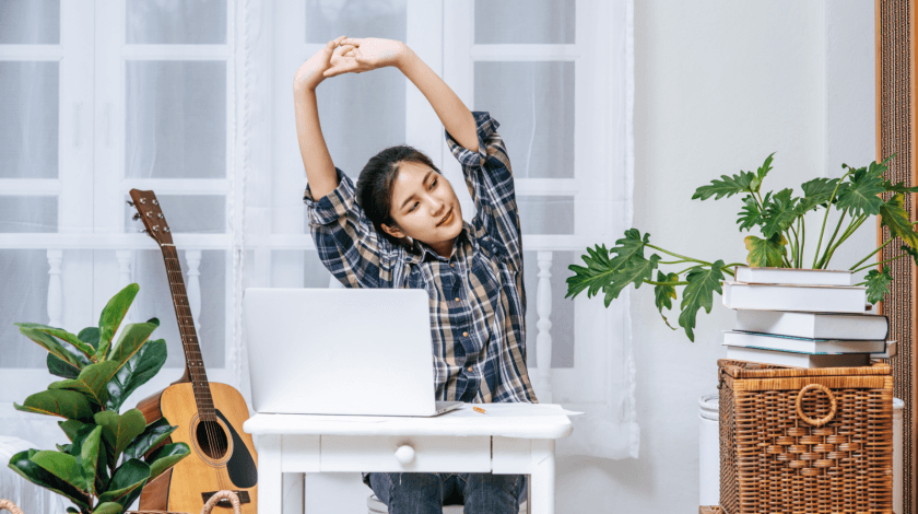 A person in a plaid shirt stretches in front of a laptop at a white desk, surrounded by indoor plants, a guitar, and stacked books, highlighting a cozy home office setting.