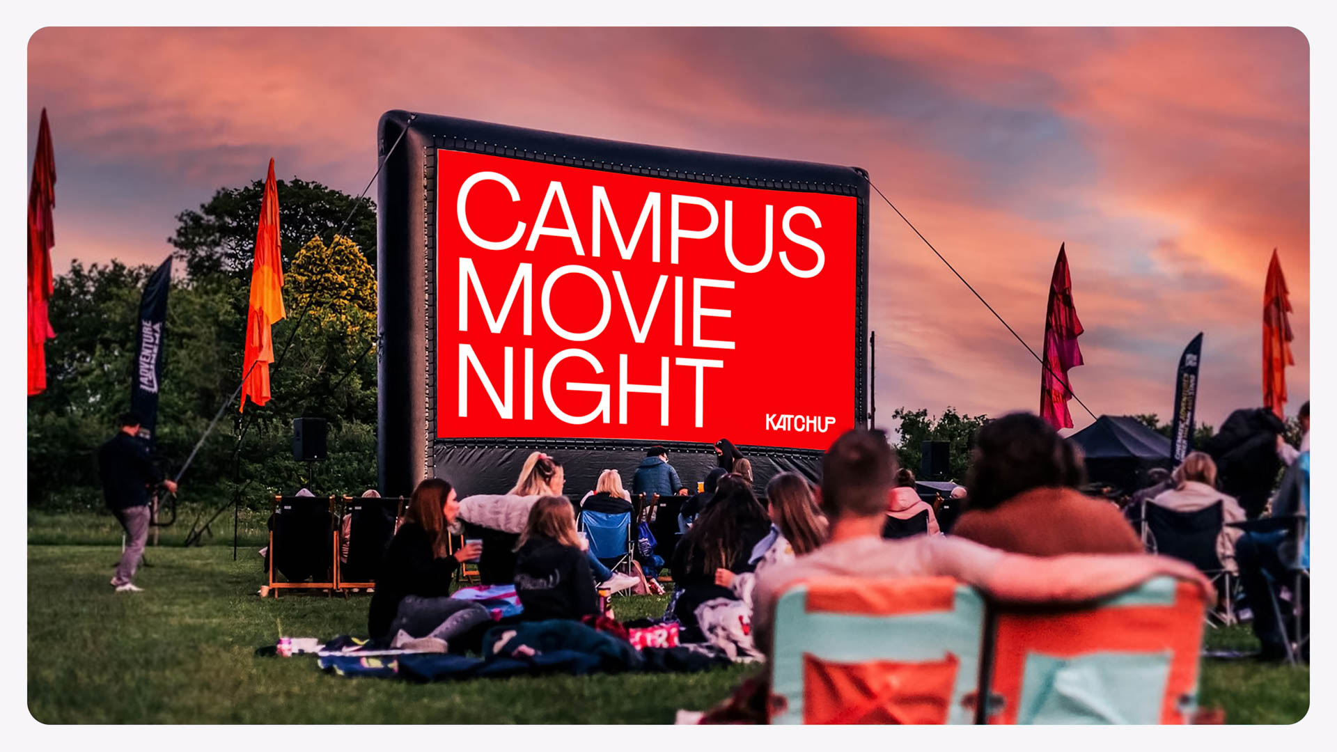 A large inflatable screen displays the words "CAMPUS MOVIE NIGHT" in white letters against a red background. The screen is mounted on a black frame and is positioned outdoors on a grassy field. Spectators are seated in lawn chairs facing the screen. Flags and banners are visible in the background. The sky above is a mix of orange and pink hues.