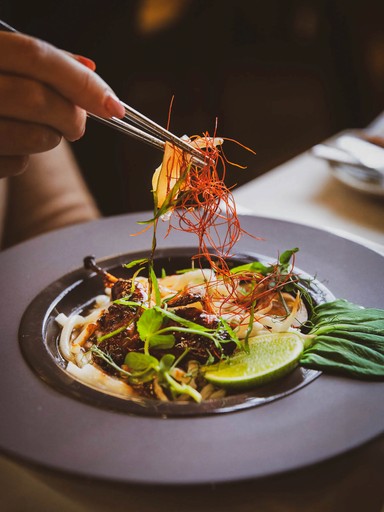 Close-up of chopsticks lifting a portion of Asian noodles with meat, garnished with herbs and chili threads, from a dark-colored bowl.