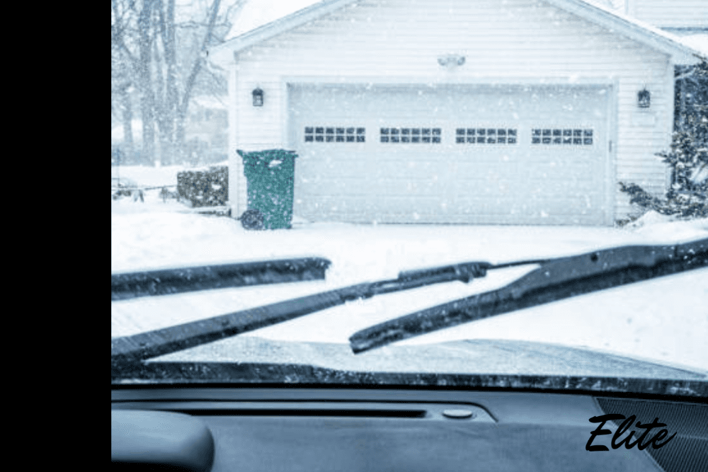Snow falling in front of a home and garage door as seen through a car windshield, showing harsh winter weather in NWA.