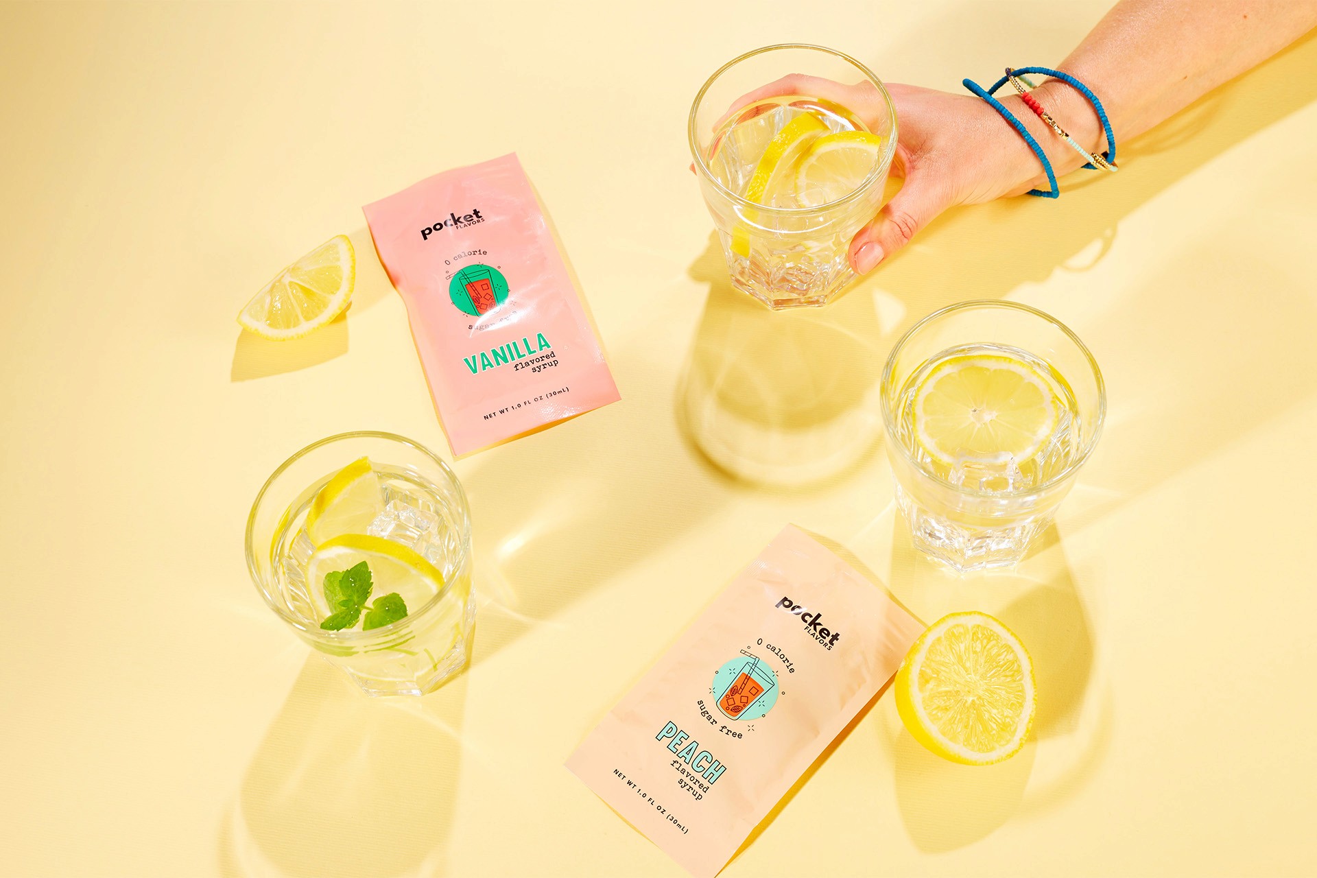 A hand wearing bracelets holds a glass of lemon water surrounded by packets of Pocket Flavors Vanilla and Peach drink mixes on a bright yellow background, with lemon slices scattered around.