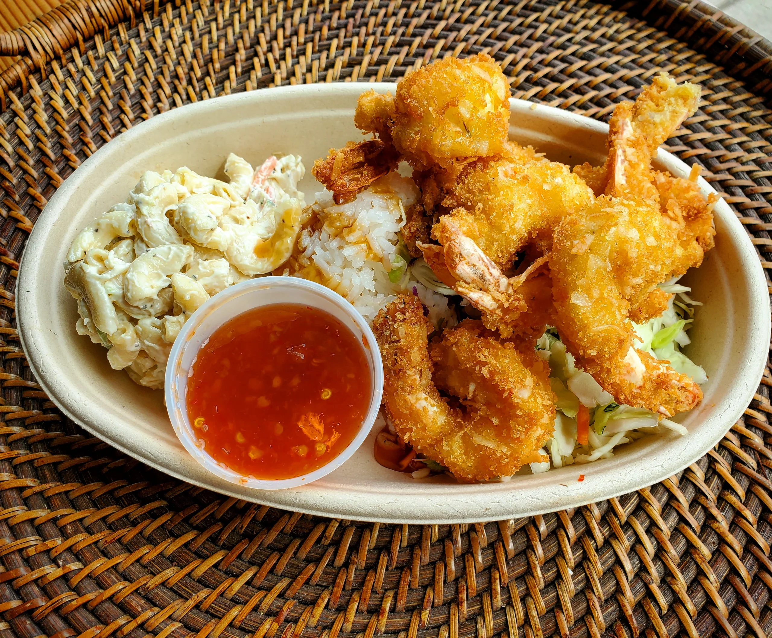 A row of golden-brown jumbo coconut shrimp served in a bowl with two scoops of rice, mac salad, and a side of red sweet chili dipping sauce.