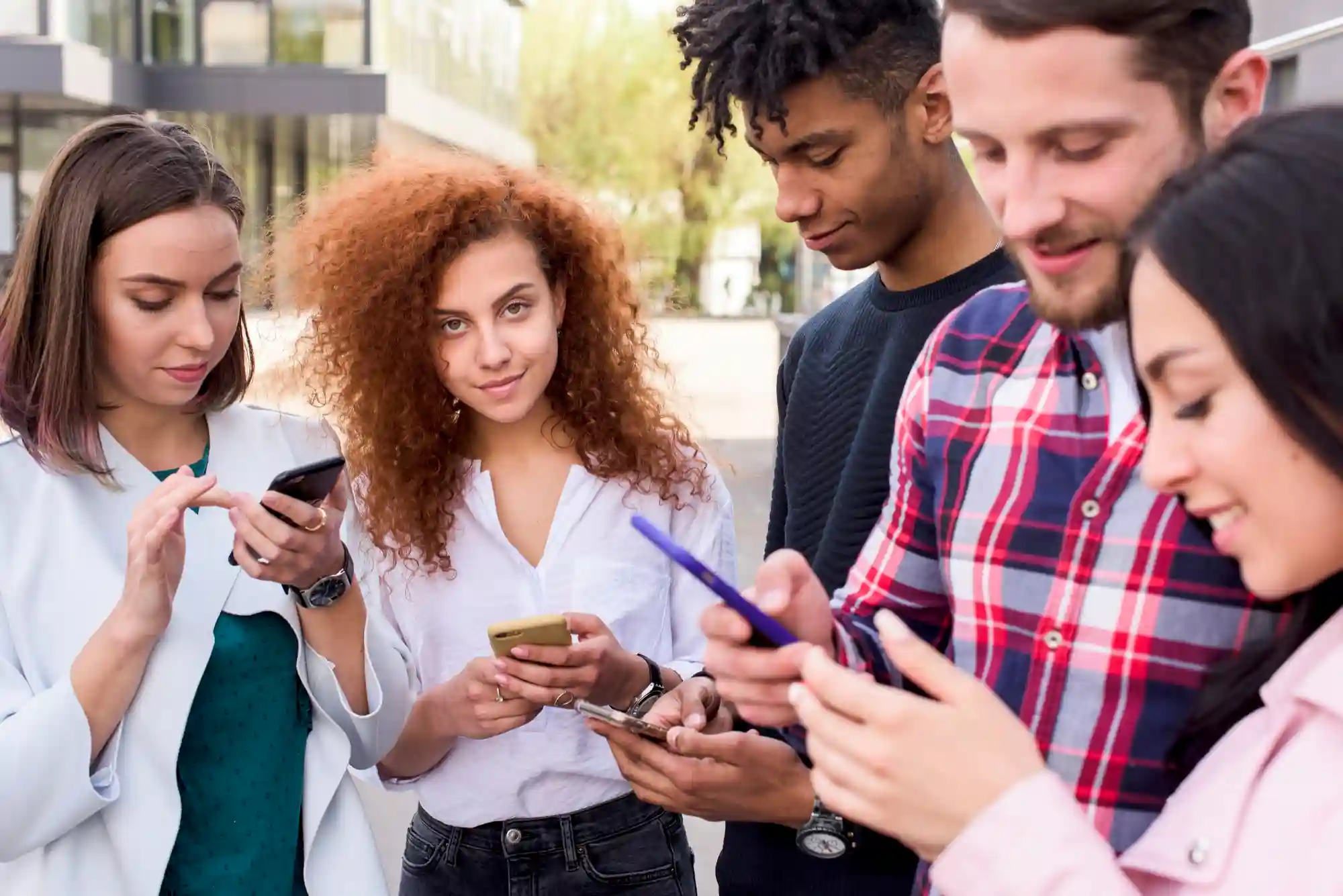 A diverse group of young friends standing together outdoors while focused on using their smartphones.