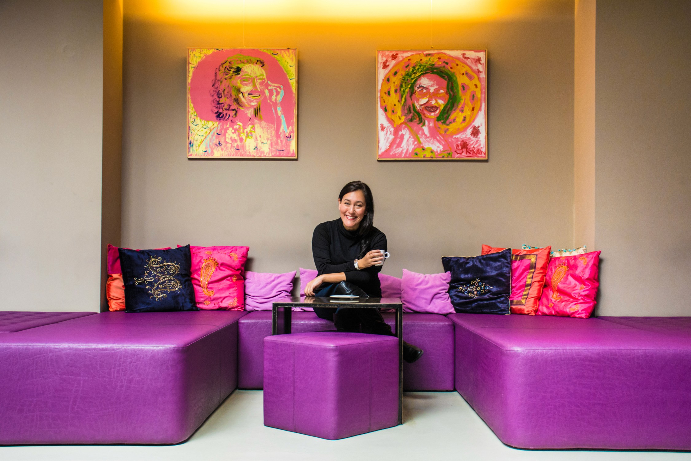 A man sits on a purple ottoman in a lounge with colorful pillows and two large abstract paintings on the wall.