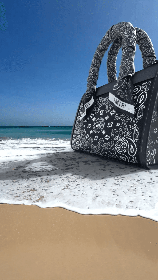A large black Amiri bag with a white paisley bandana pattern sitting on a sandy beach.