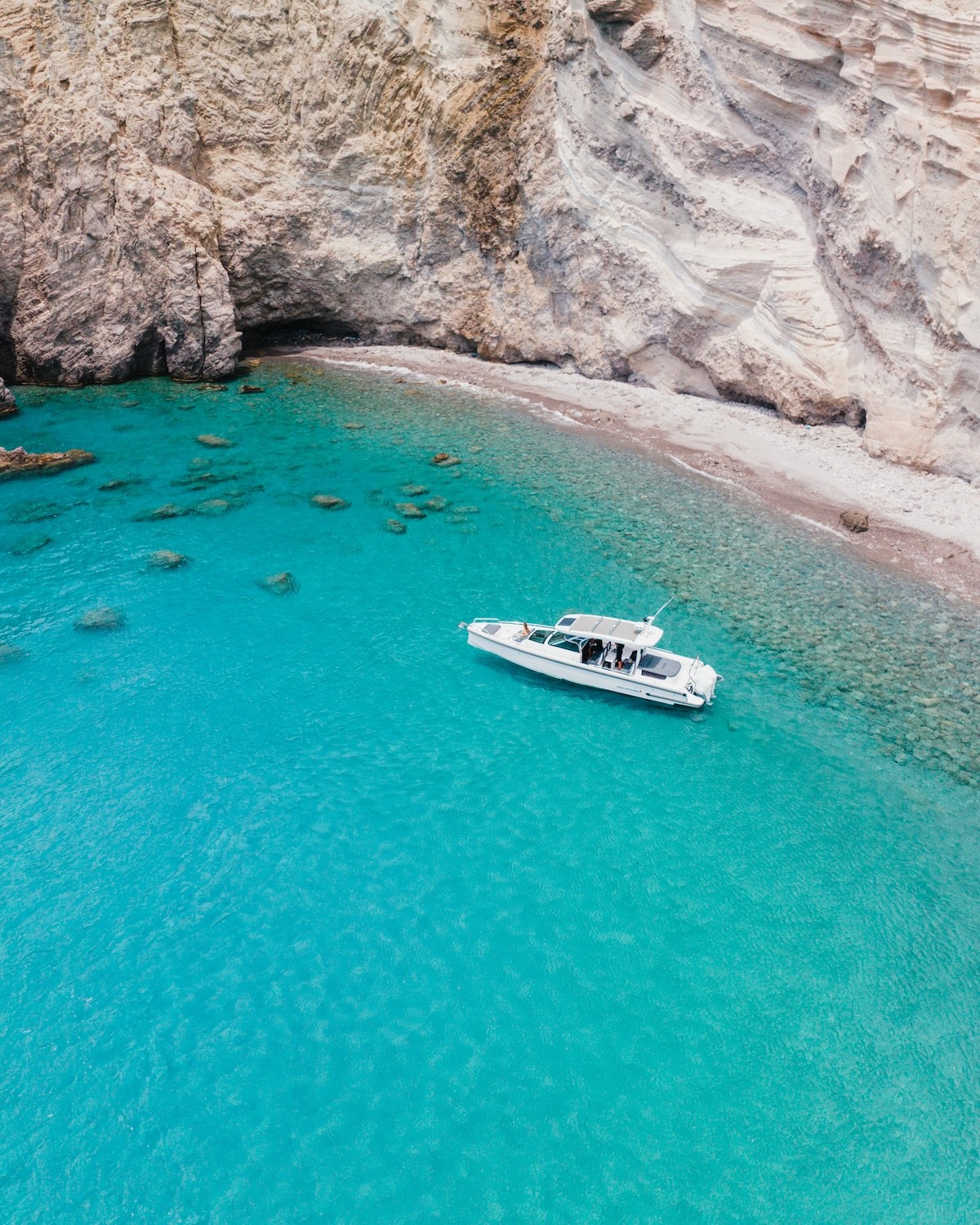 White Axopar 37 motor yacht anchored in crystal-clear turquoise waters near a rocky Cycladic coastline with mountains in the background.