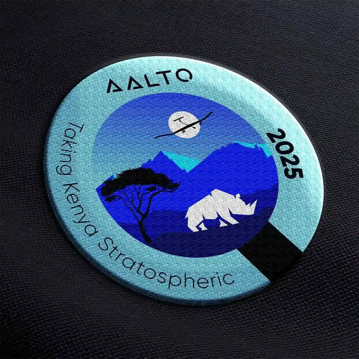 Circular mission patch for Aalto Taking Kenya Stratospheric 2025, featuring a blue layered landscape with an acacia tree, white rhinoceros, mountain silhouettes, and a stratospheric balloon against the sky