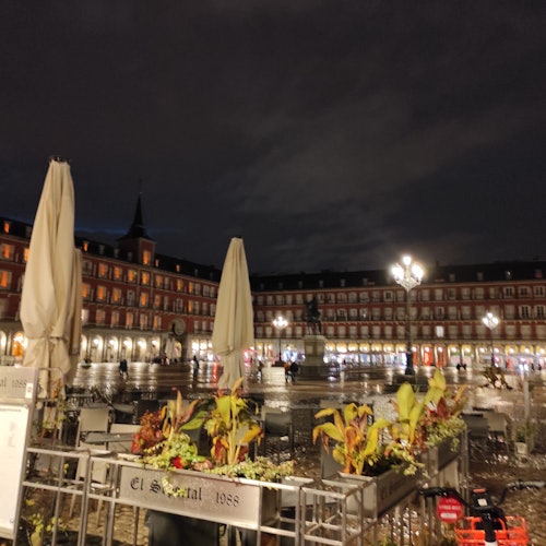 Νυχτερινή θέα μιας πλατείας με φωτισμένους φανοστάτες, κλειστές ομπρέλες, γλάστρες και ένα άγαλμα στο κέντρο, με ένα κτίριο στο παρασκήνιο.