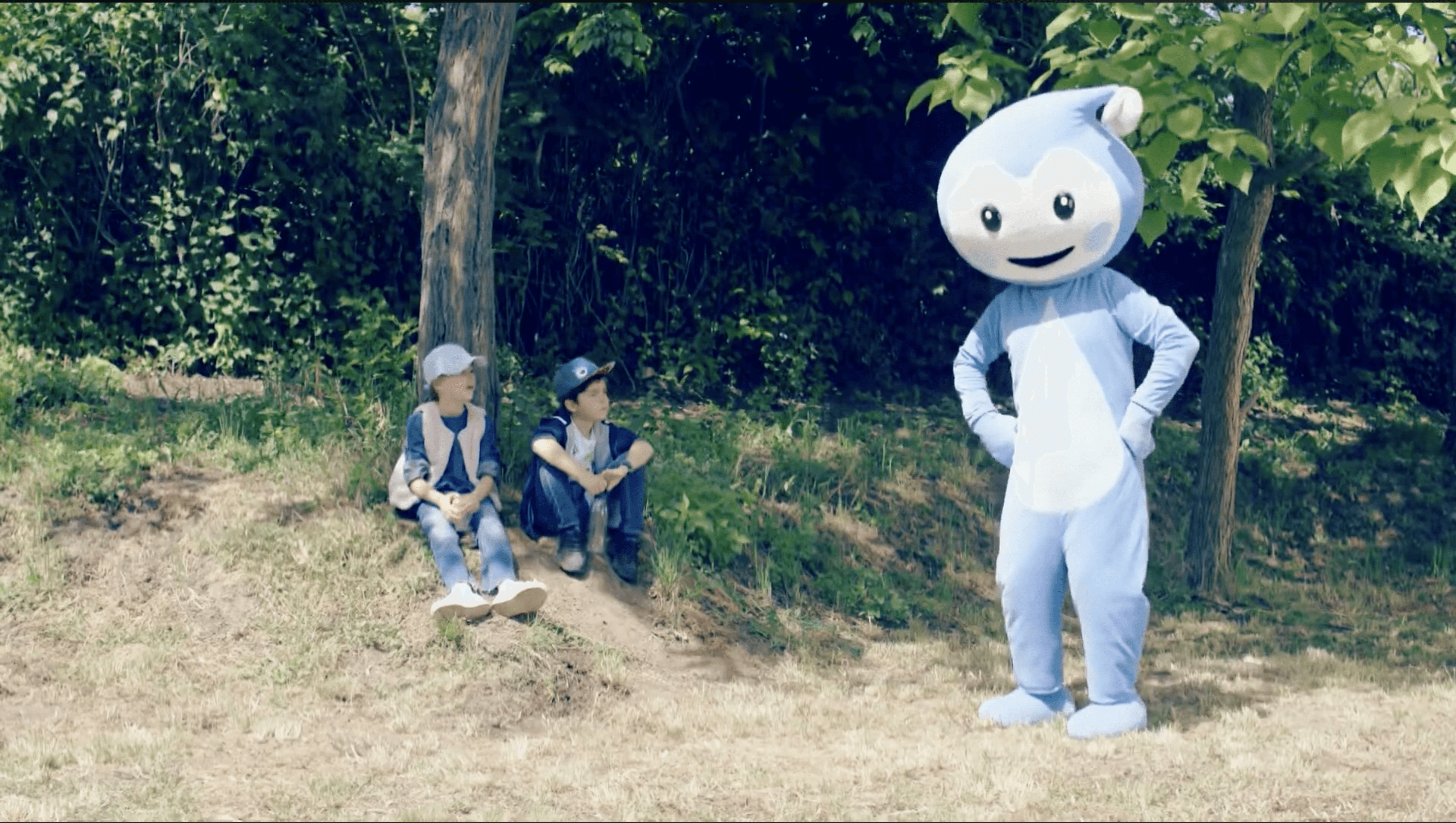 Cseppbarát kék kabalafigurája az erdő szélén táncol, a gyerekek figyelik egy fa árnyékából.